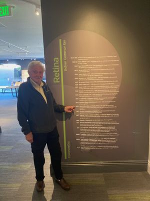 An older white mann with white hair stands in a museum gallery and points to a timelime. The museum has gray carpet and gray walls, and the green title of the timeline reads: Retina.