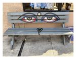 A gray park bench with a stylized painting of a pair of human eyes. The eyes are almond shaped and have black eyebrows painted over them. In between the eyes, there is a dot, and beneath the eyes, there is a swirl indicating a nose.