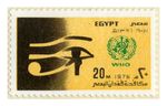 A black and yellow rectangular postal stamp with scalloped edges. On the left side of the paper there is an eye with an eyebrow with an attached swirl. On the right side at the top, it says Egypt, bellow that there is a green crest and at the bottom there are Arabic words. 
